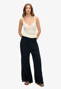 Hæklede strik tanktop i elfenben, parret med marineblå bukser med bred benvidde. Bukserne har en plisseret talje. Modellen står foran en hvid baggrund.