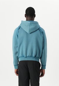 Blauer Baumwoll-Hoodie mit lockerer Passform, verstellbarem Kordelzug an der Kapuze und gerippten Bündchen. Abgenutzte Details an den Nähten verleihen Textur.