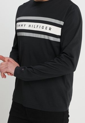 Mann trägt ein schwarzes Langarmshirt mit grauen und weißen Streifen und dem Schriftzug "Tommy Hilfiger" über der Brust, steht mit verschränkten Händen.