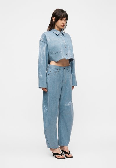 Γυναίκα που φοράει φωτεινά γαλάζιο κοντό σακάκι με παγιέτες και αντίστοιχο παντελόνι με ψηλή μέση και φαρδιά μπατζάκια, καθώς και μαύρα σανδάλια με τακούνι, στέκεται μπροστά σε λευκό φόντο.