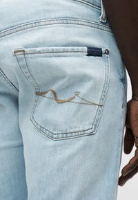 Ανοιχτό μπλε τζιν παντελόνι διαθέτει πίσω τσέπη με καμπυλωτό καφέ σχέδιο ραφής και μια μικρή ναυτική ετικέτα πάνω από την τσέπη.