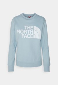 Ljusblå sweatshirt av bomull, med en stor vit logotyptext "THE NORTH FACE" och en distinkt böjd design till vänster.