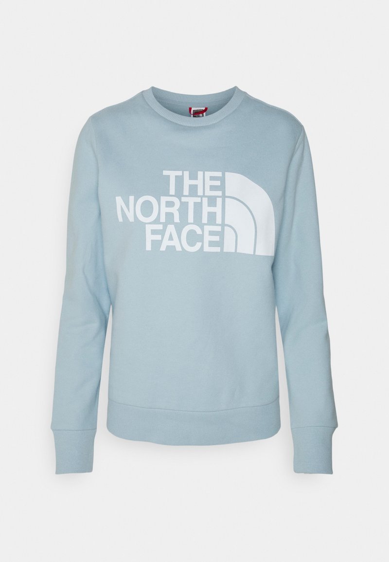 Ljusblå sweatshirt av bomull, med en stor vit logotyptext "THE NORTH FACE" och en distinkt böjd design till vänster.