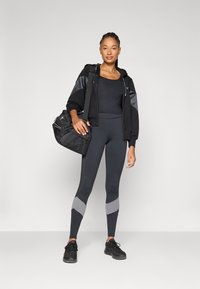 Felpa nera con zip e pannelli grigi, canotta nera, leggings con accenti grigi e scarpe da ginnastica nere; tiene una borsa da palestra nera.