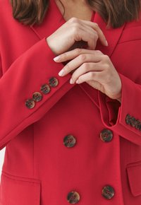 Červený blazer s přiléhavým střihem, zdobený velkými hnědými knoflíky a hladkým povrchem. Detailemi jsou prošité akcenty na rukávech.
