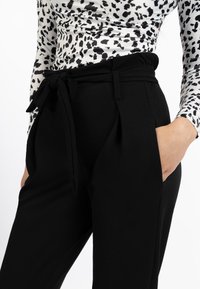 Zwarte broek met een zachte textuur, voorzien van een gestrikte taille, voorzakken en plooien. Gecombineerd met een aansluitend wit topje met een zwart patroon.