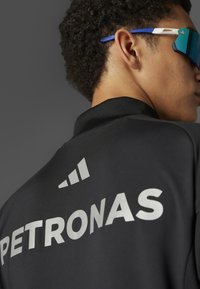 Sort jakke med "PETRONAS" i sølv, featuring høj krave og geometrisk logo. Båret med blå og hvide solbriller. Glat tekstur.