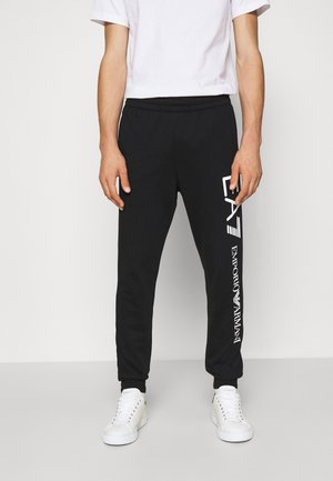Mężczyzna w czarnych spodniach dresowych z białym logo Emporio Armani na nogawce oraz białych butach sportowych na jednolitym tle.