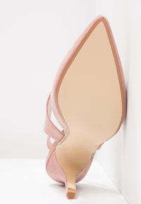 Różowy zamszowy but na wysokim obcasie z ostro zakończonym noskiem, posiadający przezroczysty pasek i gładką, beżową podeszwę. Minimalistyczny design z smukłym obcasem.