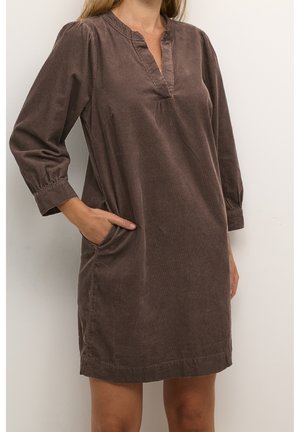 Femme portant une robe en velours côtelé marron à manches longues et col en V, main placée dans la poche latérale, debout devant un fond blanc.