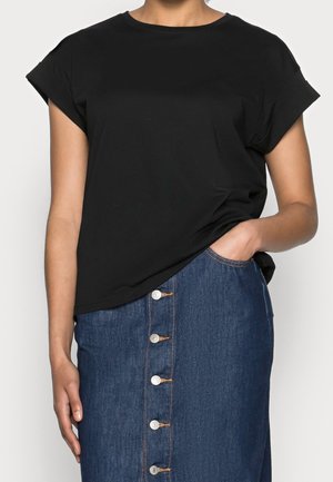 Persoon draagt een zwart shirt met korte mouwen, ingestopt in een spijkerrok met hoge taille en knopen aan de voorkant, met één hand in een zak.