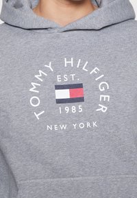 Grauer Pullover-Hoodie mit einer vorderen Tasche, versehen mit einem runden Logo, das "TOMMY HILFIGER", ein Emblem und den Text "EST. 1985 NEW YORK" zeigt.