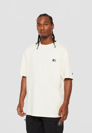 Cream oversized t-shirt with a round neckline and short sleeves. Features a small embroidered logo on the chest and a tag on the sleeve.