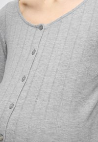 Svetlosivý sveter na gombíky s rebrovanou textúrou, ktorý má výstrih do V, šesť gombíkov vpredu a jemné vertikálne pruhy ako detail.