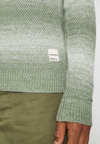 Pull en maille vert clair avec des poignets côtelés, présentant une étiquette avec le nom de la marque. Le matériau a un motif en dégradé passant à des teintes plus claires.