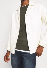 Giacca bianca con bottoni e taschino sul petto, indossata sopra un maglione verde oliva. Jeans di denim blu completano l'outfit.
