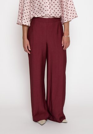 Femme portant un pantalon bordeaux taille haute à jambes larges, des chaussures beiges pointues et un chemisier rose pâle à pois rouge foncé.