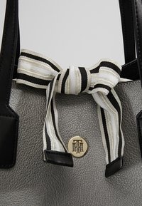 Sac à main gris texturé avec des poignées noires, orné d'un nœud en ruban rayé noir et blanc et d'un logo métallique rond avec des lettres entrelacées.