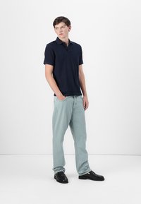 Camisa polo de manga curta azul-marinho com acabamento texturizado, combinada com jeans de perna reta azul-claro e sapatos pretos tipo slip-on sobre um fundo liso.