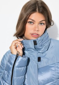 Chaqueta acolchada azul claro con cuello alto, cremallera negra y broches a presión. Presenta una textura acolchada y una tela suave y brillante.