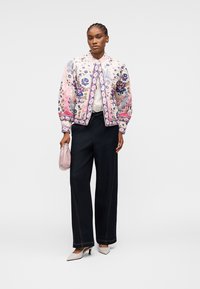 Femme portant une veste à motifs floraux, un pantalon noir à jambes larges, des talons blancs, et tenant un petit sac à main rose.