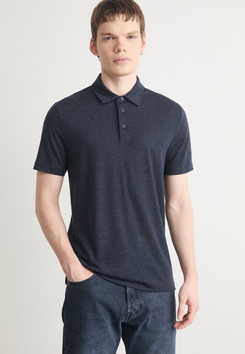 Navyblaues Poloshirt mit Kragen, kurzen Ärmeln, einer Knopfleiste mit drei Knöpfen und einer weichen, strukturierten Oberfläche. Kombiniert mit dunklen Jeans.