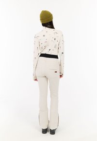 Dlhý rukáv krémová blúzka s farebnými vzormi, skombinovaná s tesnými béžovými nohavicami a čiernymi lyžiarskymi topánkami. Na hlave zelená pletená čiapka.