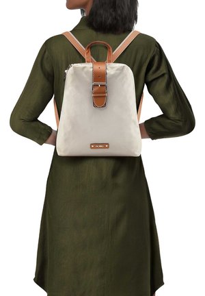 Donna che indossa un vestito verde scuro con uno zaino beige con cinturini in pelle marrone e fibbia, in piedi con le mani sui fianchi, di spalle allo spettatore.