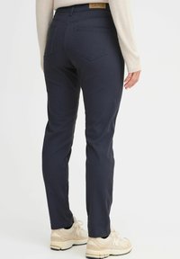Navy slim-fit pants with back pockets, a branded waistband, and a smooth texture, paired with light-colored athletic shoes.