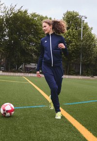 Námornícky športový teplákový komplet s bielym zipsom a logom; obuté jasne žlté futbalové topánky; kope do červeno-bieleho futbalového lopty na syntetickej trávnatej ploche.