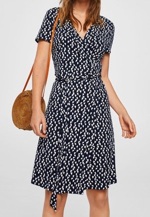 Laivastonsininen kietaisumekko lyhyillä hihoilla, jossa valkoinen lehtikuvio. Mukana solmittava vyötärö ja pyöreä olkien värinen laukku.