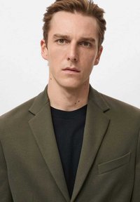 Grøn blazer med tekstureret finish, med en enkelt brystlomme, båret over en sort rundhalset skjorte, mod en ensartet lys baggrund.