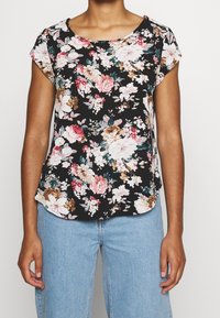 Donna che indossa una maglietta floreale nera a maniche corte con fiori rosa, bianchi e marroni abbinata a jeans azzurri, in piedi contro uno sfondo bianco.