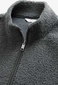 Giacca in pile grigia con colletto alto e chiusura con zip, caratterizzata da una superficie esterna testurizzata e un tessuto interno liscio. Hardware della zip in argento.