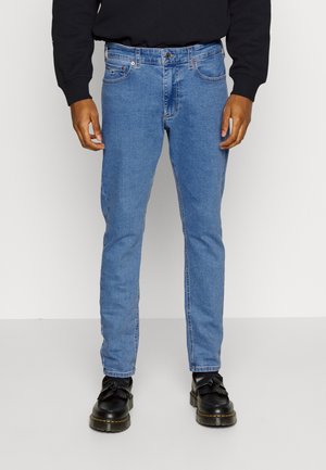 Mann trägt hellblaue Slim-Fit-Jeans, schwarze Tassel-Loafer und ein schwarzes Langarmhemd, steht vor einem schlichten weißen Hintergrund.