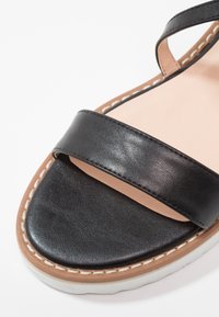 Sandale en cuir noir avec une large bride, une semelle intérieure matelassée beige et une semelle en caoutchouc blanche. Comprend des coutures contrastantes le long du bord.