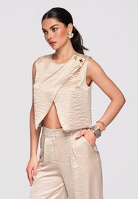 Femme portant un haut court sans manches beige texturé avec des boutons dorés et un pantalon taille haute assorti, accessoirisés avec des boucles d'oreilles florales et des bracelets.