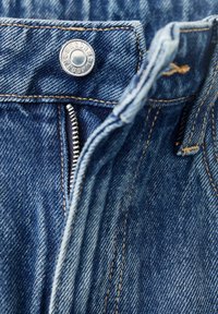 Gros plan sur un jean en denim bleu avec une fermeture éclair argentée partiellement ouverte et un bouton métallique à la taille avec des détails de couture.