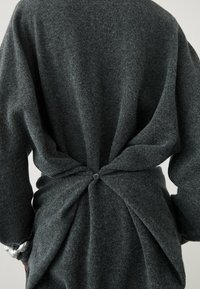 Pull gris tricoté à coupe décontractée, avec un détail torsadé dans le dos maintenu par un bouton, manches longues et texture douce.