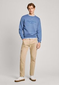 Sudadera azul claro con logo texturizado, chinos beige y zapatillas blancas. El conjunto presenta un diseño casual con cuello redondo.