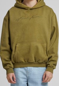 Olivově zelená mikina s kapucí, přední kapsou, a žebrovanými manžetami a lemem. Obsahuje vyšívané logo "Karl Kani" na hrudi v kontrastní barvě.