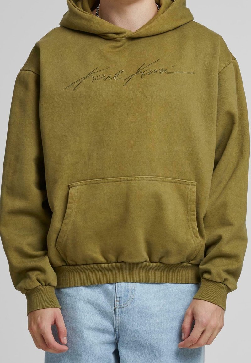 Olivově zelená mikina s kapucí, přední kapsou, a žebrovanými manžetami a lemem. Obsahuje vyšívané logo "Karl Kani" na hrudi v kontrastní barvě.