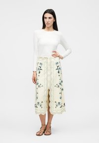 Γυναίκα που στέκεται φορώντας λευκό μακρυμάνικο τοπ, κρεμ midi φούστα με floral κέντημα και κυματιστές άκρες, και καφέ σανδάλια με λουράκια.