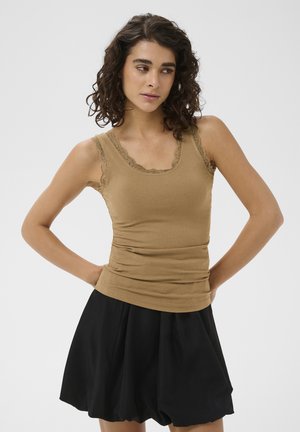 Femme aux cheveux bouclés portant un haut côtelé beige sans manches avec bordure en dentelle et une jupe évasée noire, debout les mains sur les hanches.