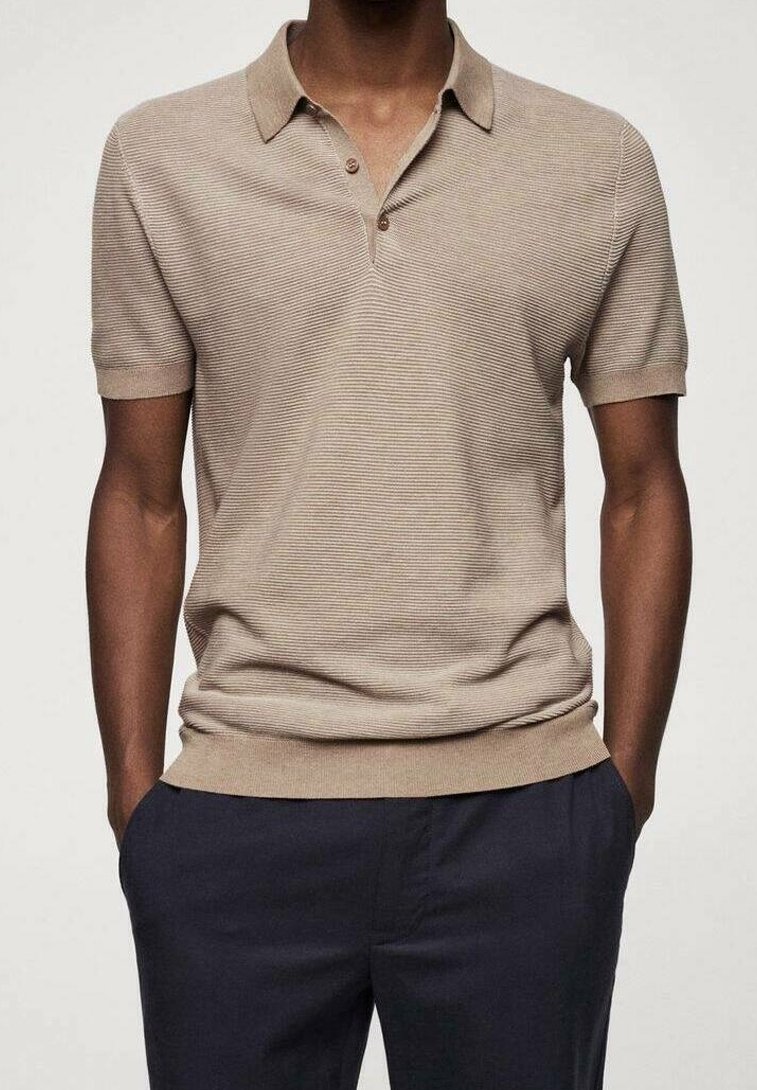 Homme portant un polo beige texturé à manches courtes avec deux boutons, associé à un pantalon bleu marine foncé, mains dans les poches.