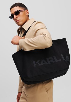 Crna platnena torba s mat završnom obradom, s velikim tekstom "KARL LAGERFELD" u kontrastnoj boji. Podesiva naramenica.