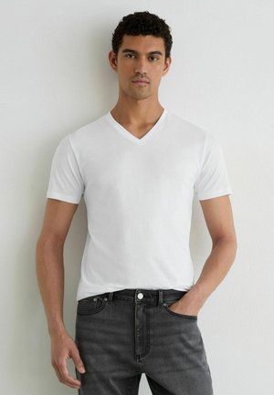 Jonge man met krullend haar die een effen witte T-shirt met V-hals en zwarte spijkerbroek draagt, staand met één hand in de zak tegen een lichte muur.