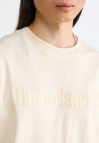 Krämfärgad bomulls-T-shirt med mjuk, broderad "Timberland"-text och logotyp. Rundad halsringning med ribbad kant. Mjuk textur.