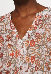 Top con estampado floral en tejido ligero que presenta un escote en V y un diseño con rojos, verdes y amarillos sobre un fondo blanco.