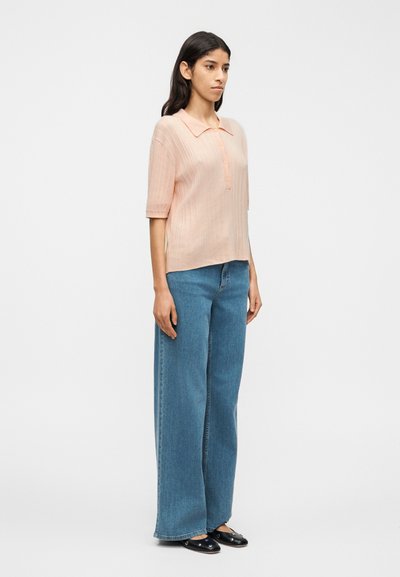 Ροζ ανοιχτόχρωμο πουκάμισο με κοντά μανίκια και γιακά, με κάθετες ρίγες, συνδυασμένο με φαρδιά μπλε τζιν παντελόνια και μαύρα παπούτσια slip-on.